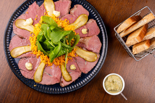 Top-down close-up of an appetizer plate featuring pastrami, roast beef, arugula, and toast on a wooden table.