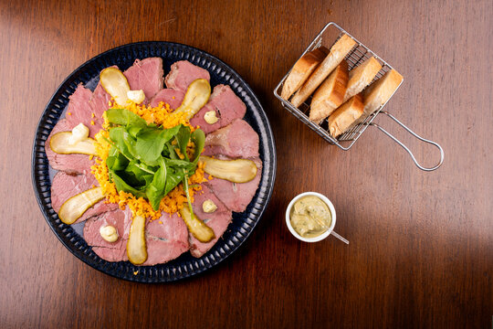Top-down view of a pastrami and roast beef appetizer with arugula and toast on a wooden table.