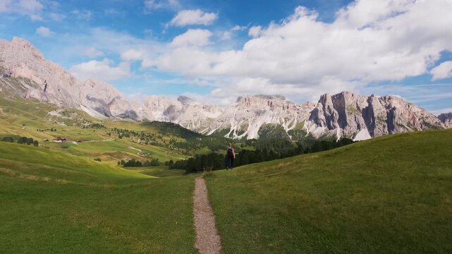 Slow pull back on meadow path as hiker walks toward forest and valley under jagged peaks in sunny daylight, Dolomites, South Tyrol, Alps, Italy