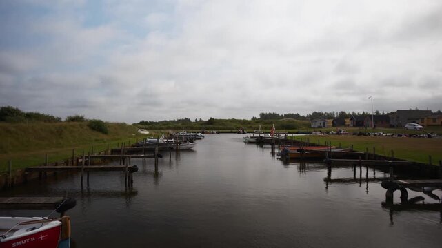 Static hold along a calm marina channel with wooden jetties and moored boats between grass banks in overcast light, Scandinavia, Denmark