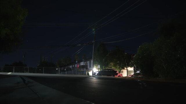 Car driving along a dark urban street late at night, passing an intersection among residential buildings, trees, power lines and dim lighting