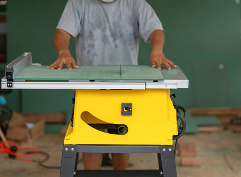 A carpenter adjusting and guiding a green board on a yellow electric table saw in a woodworking workshop.