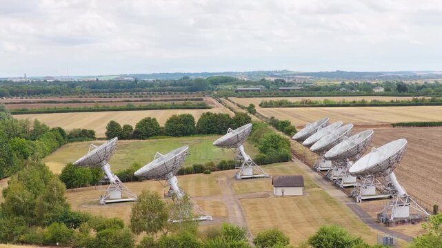 Large white satellite dishes slowly move and pivot toward the sky to capture signals across a wide grassy field during a bright day under a cloudy horizon.