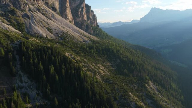 Aerial push and yaw along sunlit limestone cliffs above dense pine forest overlooking misty valley in morning, Dolomites, South Tyrol, Alps, Italy