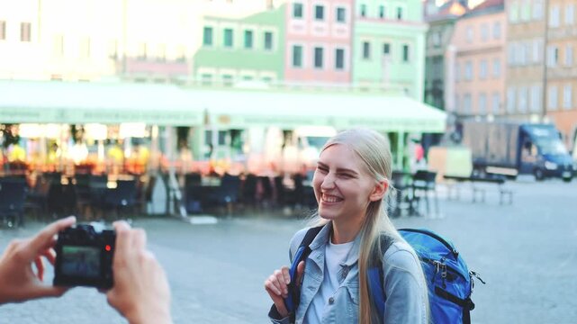 A man takes a photo of a smiling blonde woman with a blue backpack in a colorful city market square with outdoor seating and pastel buildings during daytime.