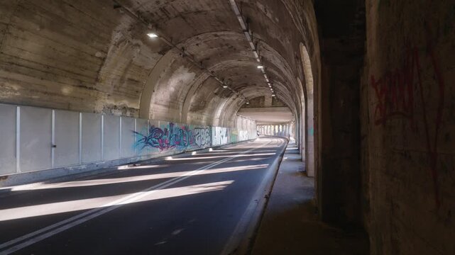 Static view down empty arched road tunnel with striped sunlight falling on asphalt and graffiti walls, Lago Maggiore, Piedmont, Lombardy, Italian lakes, Italy