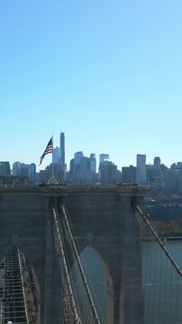 Vertical video: Aerial slide and pan footage of US flag waving on top of Brooklyn Bridge. Parallax effect with modern high rise downtown buildings. New York City, USA