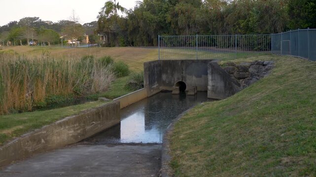 Slow motion landscape of a public concrete stormwater drainage outlet with baffle blocks and culvert channel leading to a grassy retention waterway park basin in rural town suburbs of NSW Australia