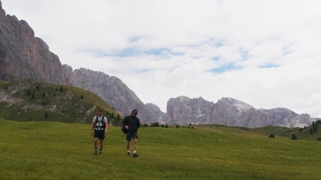Static shot of two hikers walking toward camera across green alpine meadow beneath limestone cliffs in daylight, Dolomites, South Tyrol, Alps, Italy