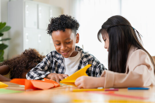 Multiethnic kids doing origami paper craft in classroom, diverse school children folding paper in kindergarten class, cute african american boy and asian girl learning arts and crafts at school desk