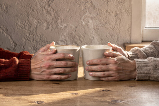 Two people holding steaming ceramic mugs on rustic wooden table