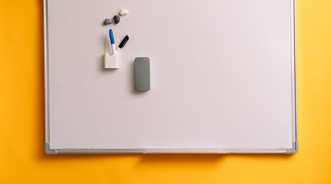 Cropped blank whiteboard with two markers on a yellow wall with a gray eraser.
