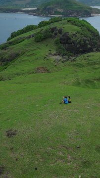 Couple sitting grassy cliff overlooking ocean, aerial drone perspective captures turquoise bay, rocky shoreline and sweeping green hillside backpacks beside pair, Lombok, vertical video