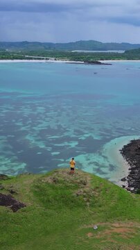 Man standing on cliff overlooking turquoise bay, solo traveler wearing yellow shirt gazing over coral reef and shallow lagoon, aerial view reveals clear water, distant islands and cloudy,Lombok