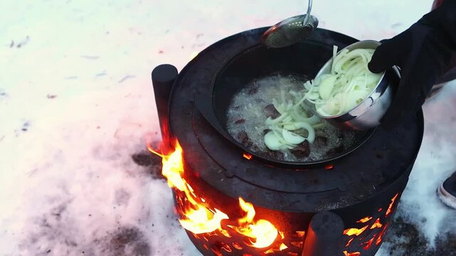 Close-up of onions being poured into a pan during outdoor winter cooking. Meat simmers over open flames while thick steam rises into the cold snowy air.