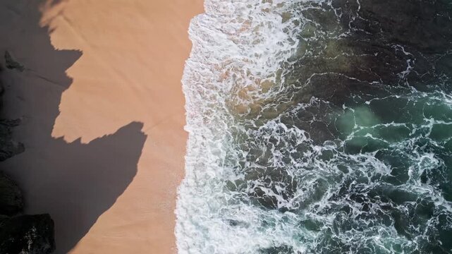 Aerial bali coastline rocky beach waves washing over golden sand and dark volcanic rocks, turquoise water swirling into white foam, dramatic shadows from cliffs, soothing rhythmic, cinematic video