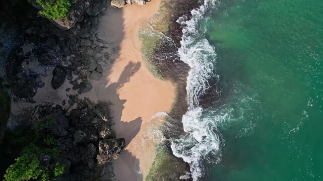Aerial bali coastline rocky beach waves washing over golden sand and dark volcanic rocks, turquoise water swirling into white foam, dramatic shadows from cliffs, soothing rhythmic, cinematic video