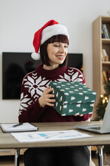 A smiling Caucasian woman wearing a Santa hat and a festive sweater holds a Christmas gift box while sitting at a desk with a laptop © Sofiia Shunkina