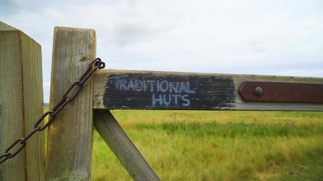 weathered timber gate blocks the entrance to a lush pastoral field. The wood features a charming, hand-painted "Traditional Huts" sign, evoking a peaceful, rural vacation atmosphere.