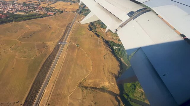 A cinematic aerial perspective from a passenger window showing an airplane wing soaring over golden fields, a river, and a highway during a sunset flight.