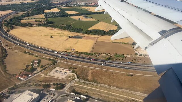 A passenger perspective from an airplane window showing the wing over a busy highway with traffic, agricultural fields, and rural infrastructure during a bright day.