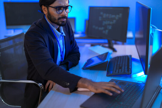 A bearded software developer in a dark suit and glasses typing program code on the keyboard, at a multi-monitor workstation in a blue-lit technology office; over-the-shoulder angle.