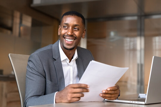 Portrait of smiling Black businessman sits at office desk, checking document, working on report, reading business proposal, manage corporate documentation, reviewing information with positive results