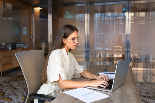 Caucasian adult businesswoman sitting at office desk typing on laptop, working on corporate tasks, managing digital communication, preparing business report, responding on email, reviewing information