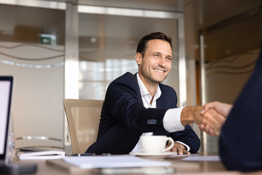 Smiling Caucasian businessman shaking hands with partner starting business meeting, making agreement, concluding partnership at desk in corporate office. Professional communication, deal, recruitment