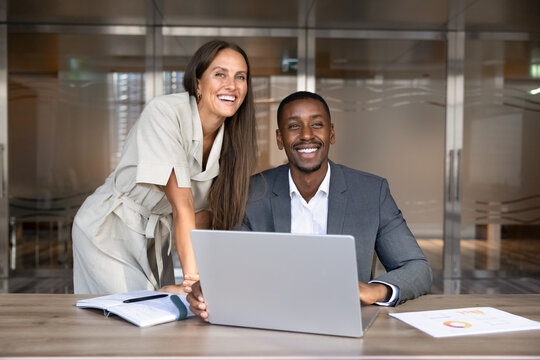 Smiling multiethnic colleagues gather at laptop in office to review project plan, develop marketing strategy, joint business task discussion, portrait. Teamwork, successful corporate collaboration