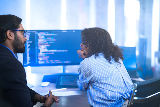A male team lead leaning over a seated female developer explaining a fix on the monitor, to point at code on her monitor in a blue-lit office; over-the-shoulder angle of software frame6489.