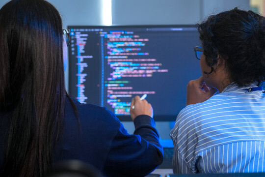 Two engineers seen from behind analysing the program on multiple displays, facing a wall of monitors filled with source code; over-the-shoulder angle of software development and dedication.