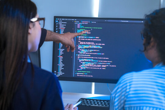 Two engineers seen from behind watching data scroll across the monitors, facing a wall of monitors filled with source code; close-up of software development and solution in a blue-lit.