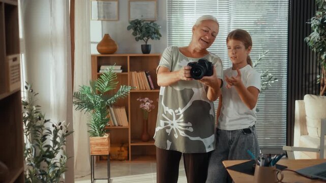 Grandmother teaches her granddaughter how to use a DSLR camera in the living room, sharing photography skills, technology knowledge and a warm intergenerational family moment at home.