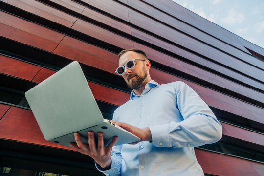 Businessman in stylish sunglasses working on laptop outside office, remote work