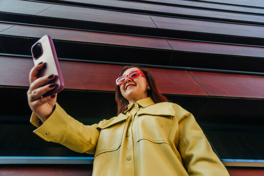 Redhead white woman in yellow shirt and bright glasses taking selfie with phone