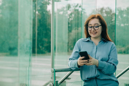Redhead businesswoman in blue suit using phone app, glasses, looking at display