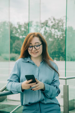 Redhead businesswoman in blue suit using phone app, glasses, looking at display