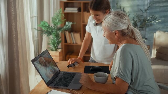 Happy grandmother invites her granddaughter to join a family video call on a laptop, sharing laughter, joy and a warm intergenerational moment in the living room.