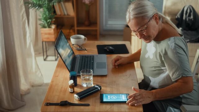 Mature woman works on a laptop at her home desk when a medication reminder alarm appears on her smartphone, notifying her that it is time to take her scheduled medicine or vitamins.