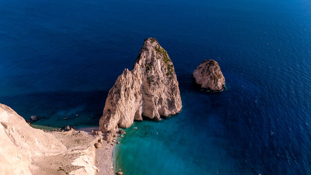 Aerial view of two limestone sea stacks rising from deep blue sea beside a white cliff and turquoise cove, Myzithres, Zakynthos, Greece