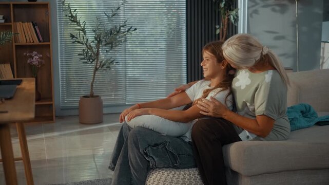 Young granddaughter sits beside her grandmother and receives a warm hug as they smile together in the living room, showing affection, comfort and close family bonding at home.