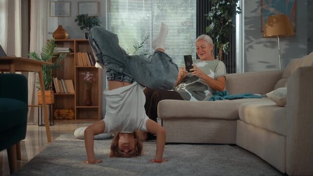 Young girl performs a headstand on the living room floor while her grandmother records her with a smartphone, sharing a playful and supportive family moment at home.