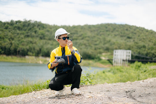 Professional engineer wearing a hard hat operates a drone remote while talking on a walkie-talkie. Outdoor site inspection near a scenic lake