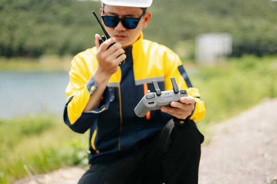 Professional engineer wearing a hard hat operates a drone remote while talking on a walkie-talkie. Outdoor site inspection near a scenic lake