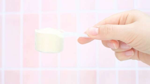 Female hand holding measuring scoop with protein powder on pink tile background side view.