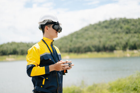Male engineer in safety gear uses FPV goggles and a remote to pilot a drone. Conducting a high-tech aerial survey near a beautiful natural lake