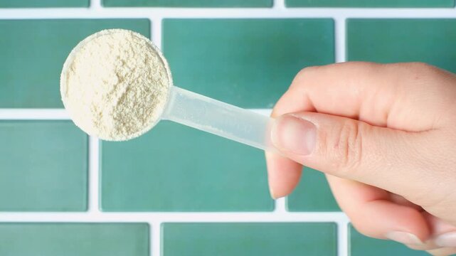 Female hand holding measuring scoop with protein powder on blue tile background top view.