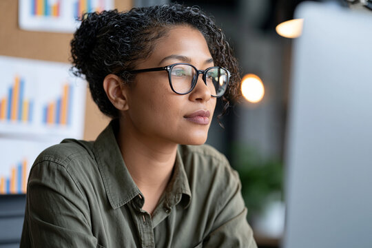 Professional black woman wearing glasses, thinking and concentrating while working on a computer in a modern office, developing business strategy and analyzing data, copy space