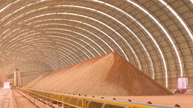 Industrial factory complex with large piles of crushed stone aggregate material like limestone pouring from conveyor belts, before being heated to extremely high temperatures to produce cement.